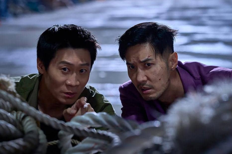 Dua pria bersembunyi di balik tumpukan tali dalam adegan tegang film Husbands in Action, dengan ekspresi waspada di wajah mereka.