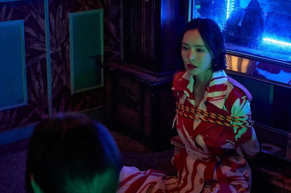 Seorang wanita mengenakan mantel bermotif merah putih duduk di lantai dengan tangan terikat tali di ruangan bercahaya neon biru.