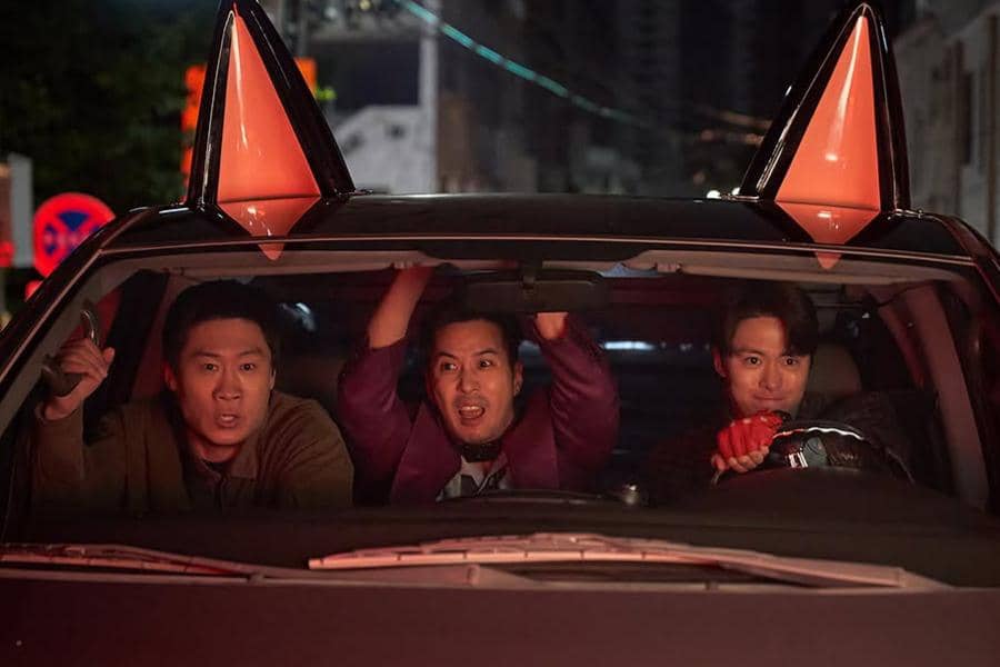 Tiga pria duduk di dalam mobil pada malam hari dengan ekspresi tegang dan lucu, adegan dari film Husbands in Action.