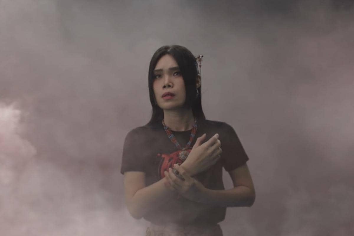 Seorang perempuan berdiri di tengah kabut tebal dengan ekspresi tegang, mengenakan kaus hitam dan kalung berwarna merah biru.