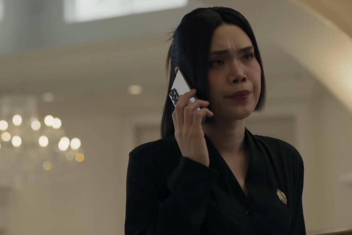 Seorang wanita berambut hitam mengenakan pakaian hitam sedang berbicara di telepon dengan ekspresi serius di dalam ruangan bercahaya lembut.