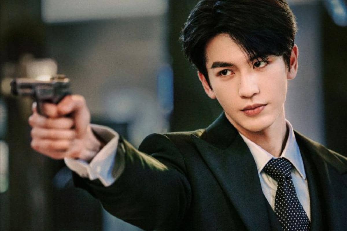 Seorang pria berpakaian jas hitam memegang pistol dan mengarahkannya ke depan dengan ekspresi serius dalam adegan drama Overdo.