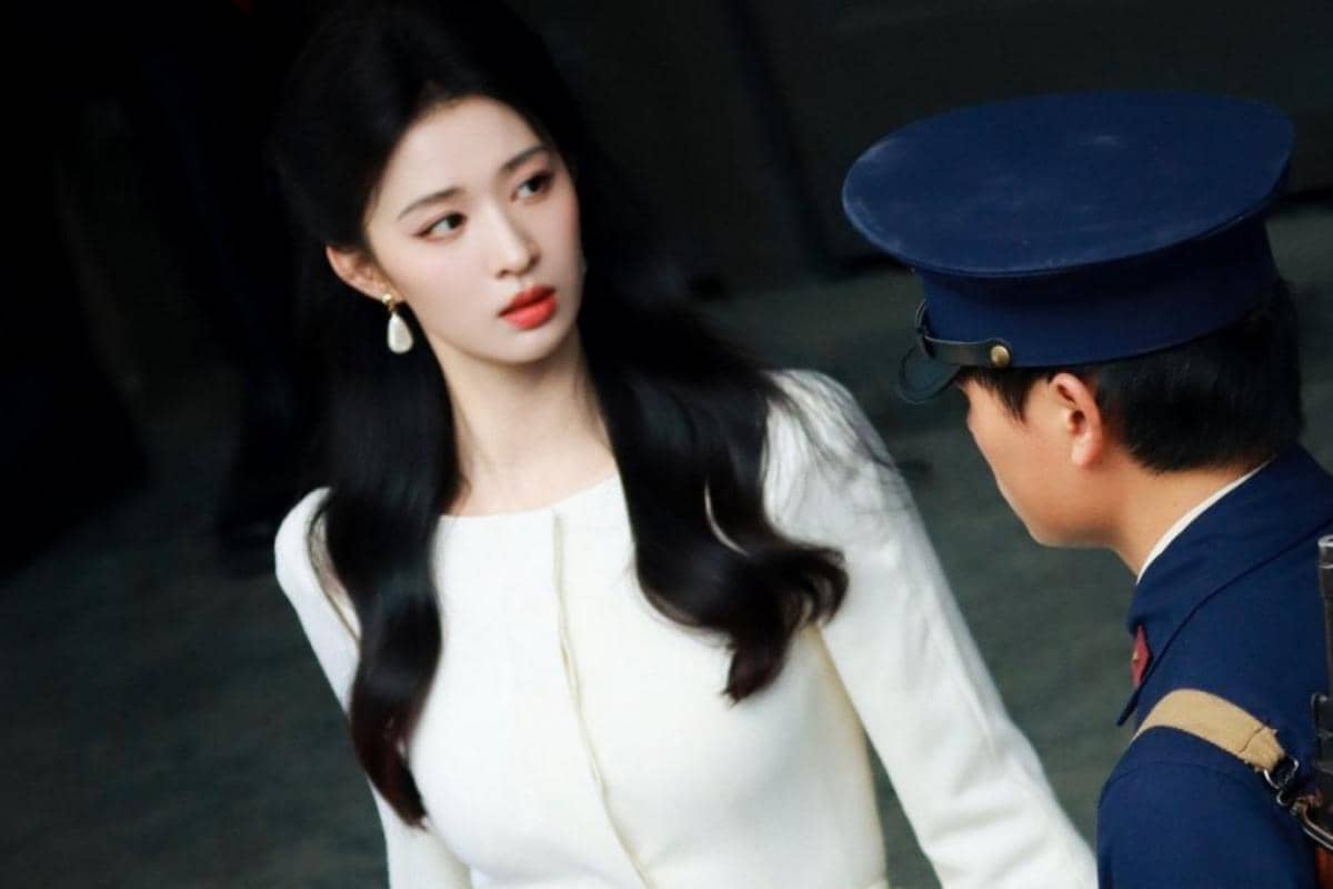 Seorang wanita berambut panjang mengenakan gaun putih berbicara dengan pria berseragam biru dalam adegan drama Overdo.