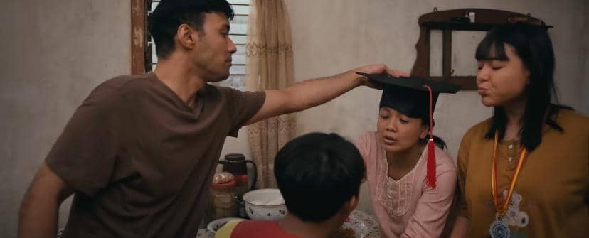 Seorang pria menyentuh kepala wanita yang mengenakan topi wisuda di dalam ruangan sederhana bersama dua orang lainnya.