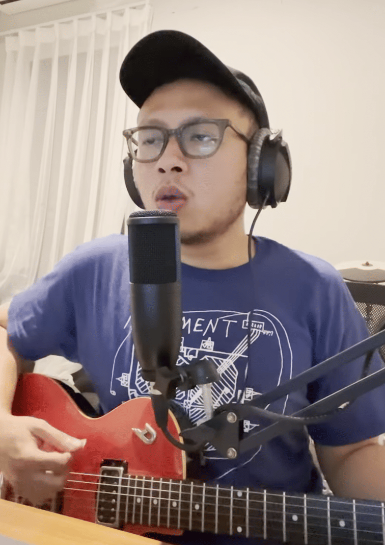Seorang pria mengenakan kacamata dan topi sedang bernyanyi sambil memainkan gitar listrik merah di depan mikrofon dan memakai headphone.