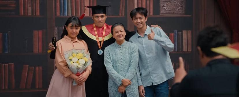 Empat orang berpose bersama, salah satunya mengenakan toga wisuda dan medali, sementara yang lain tersenyum di depan latar rak buku.