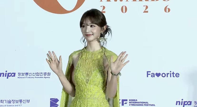 Seorang wanita mengenakan gaun hijau cerah melambaikan tangan di karpet merah acara Global OTT Awards 2026 di Korea.