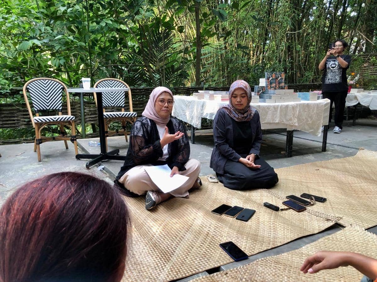 Dua perempuan duduk bersila di atas tikar dalam acara peluncuran buku di ruang terbuka dengan latar pepohonan hijau.