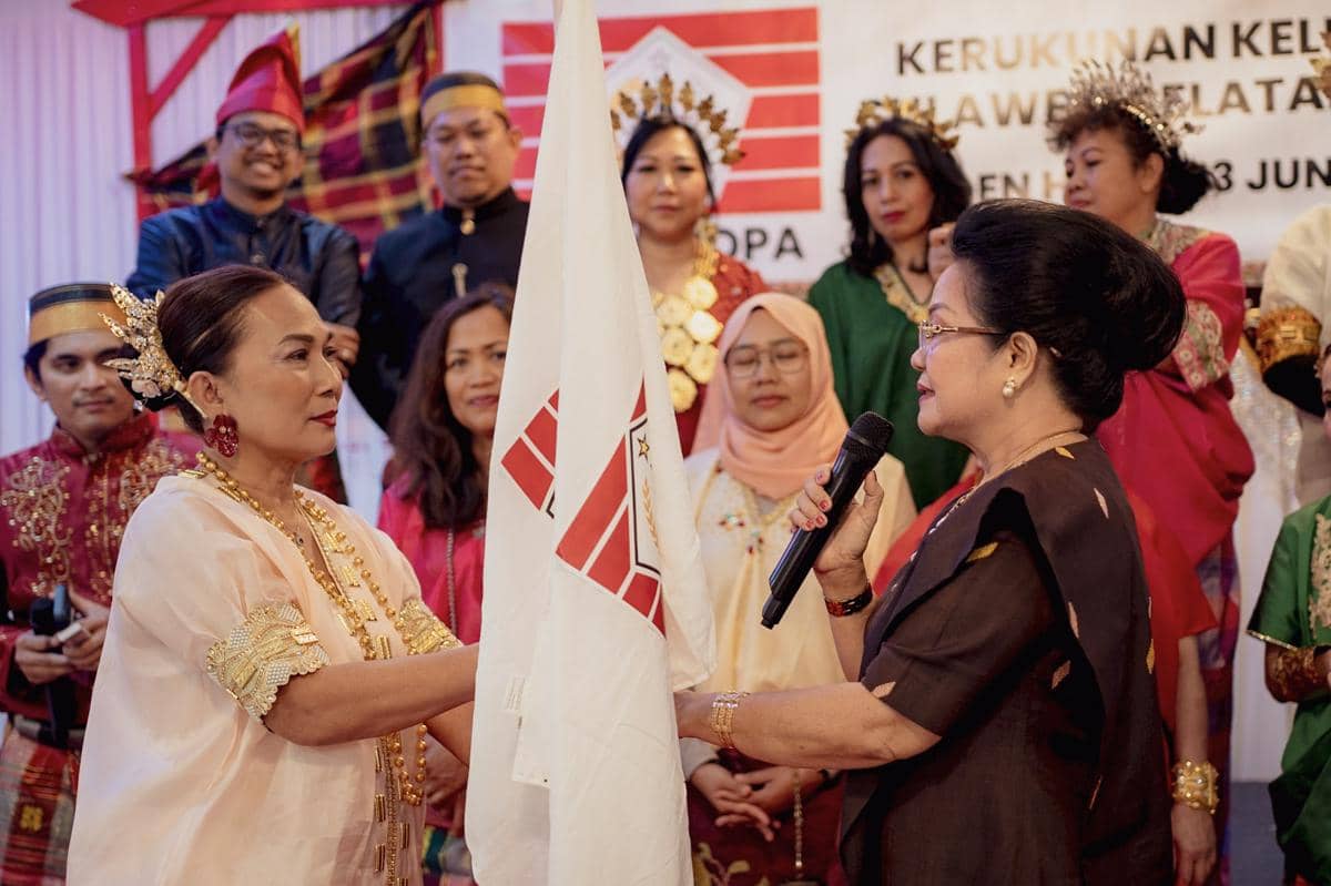 Seorang perempuan menerima bendera organisasi dari perempuan lain dalam acara pelantikan KKSS Eropa, disaksikan peserta berpakaian adat Bugis-Makassar.
