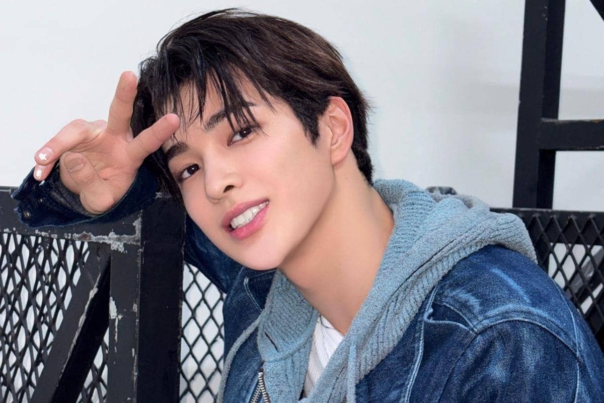 Seorang pria muda mengenakan jaket denim dan hoodie abu-abu tersenyum sambil mengangkat dua jari membentuk tanda damai.