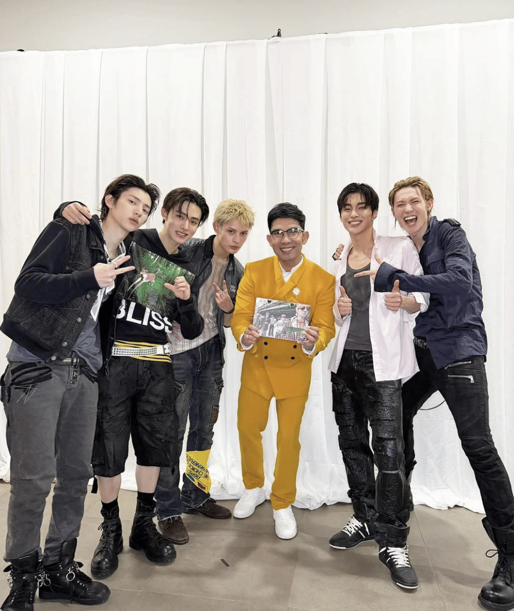 Seorang pria mengenakan setelan kuning berfoto bersama enam pria muda berpakaian kasual sambil memegang album bertanda tangan.