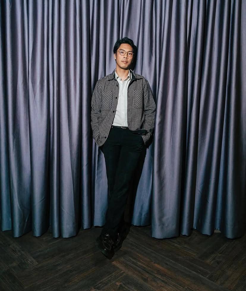 Seorang pria berdiri santai dengan tangan di saku di depan tirai berwarna abu-abu, mengenakan kemeja dan jaket bermotif kotak.