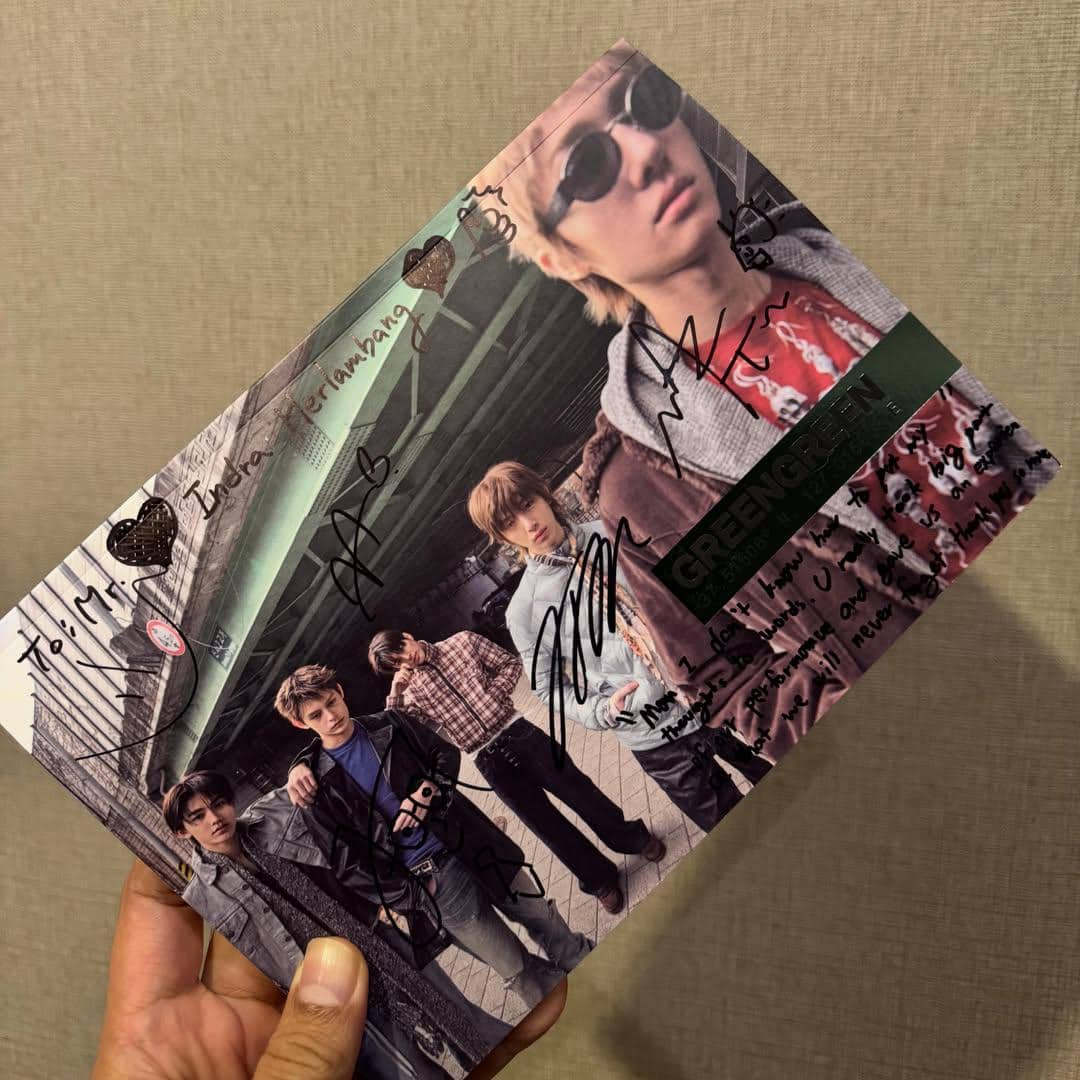 Seseorang memegang album bertanda tangan dengan foto lima anggota grup musik di bawah jembatan, berlatar suasana perkotaan.