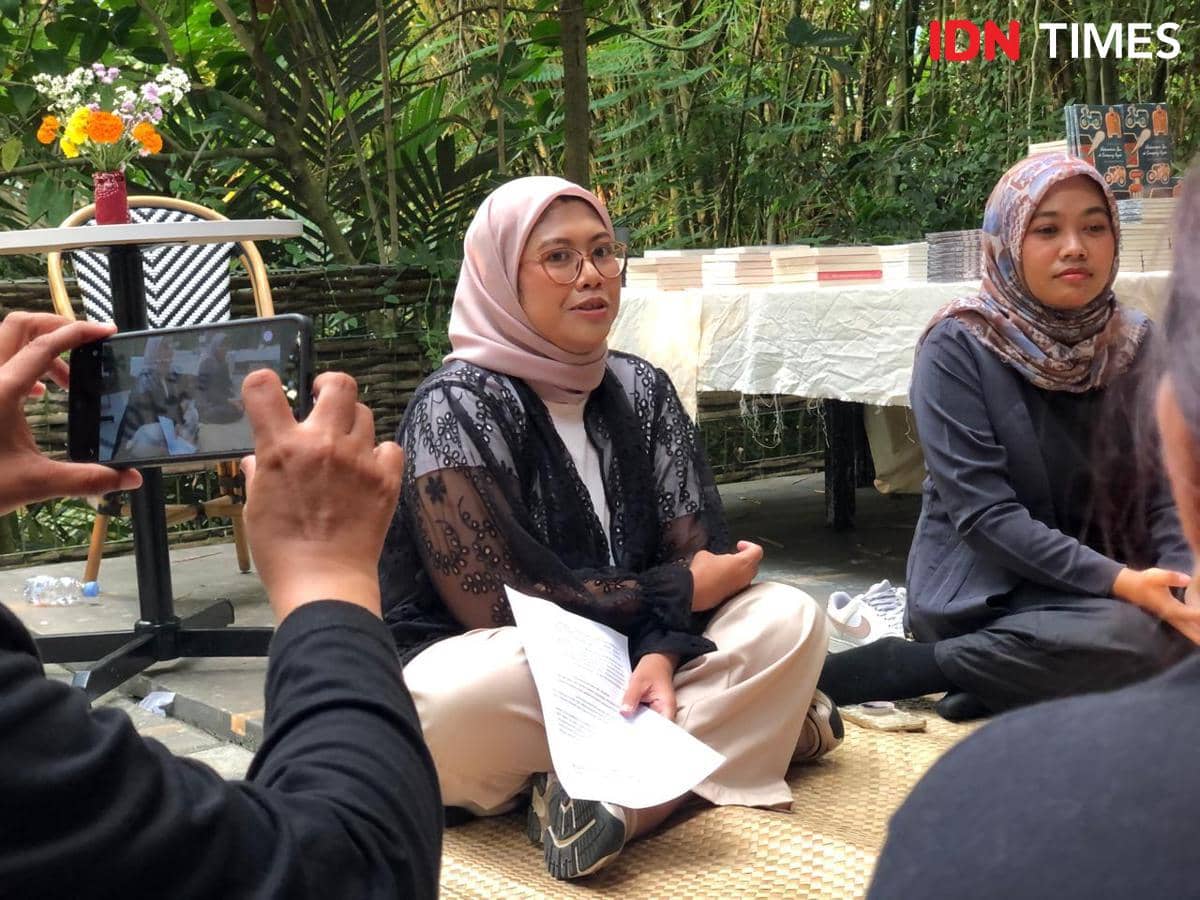 Seorang perempuan berhijab duduk di atas tikar sambil memegang kertas dalam acara peluncuran buku di ruang terbuka dengan latar tumpukan buku.