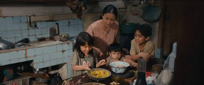 Seorang ibu bersama tiga anaknya sedang menyiapkan makanan di dapur sederhana dengan peralatan masak dan bahan makanan di meja.