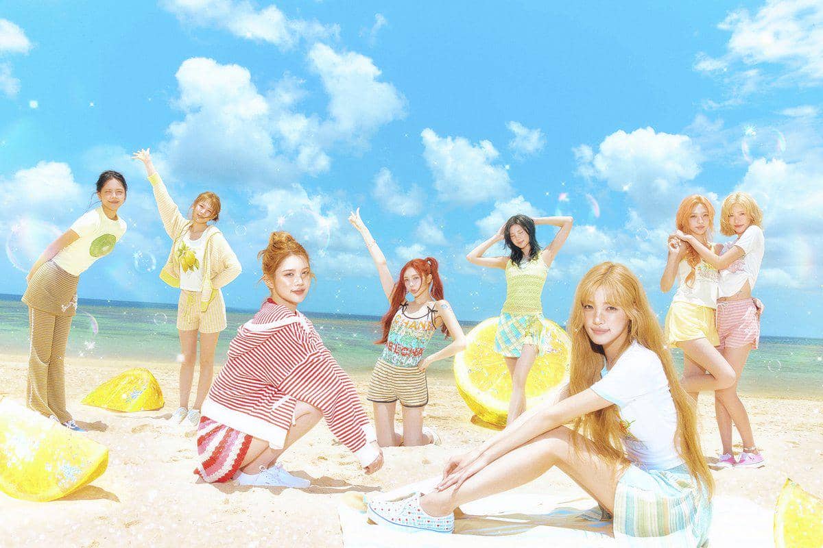 Tujuh anggota Hearts2Hearts berpose ceria di pantai dengan latar langit biru dan potongan lemon besar untuk mini album Lemon Tang.
