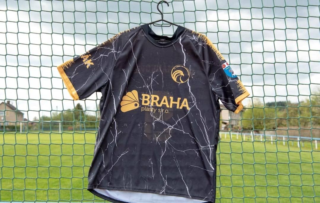 Jersey sepak bola berwarna hitam dengan motif garis putih dan aksen kuning tergantung di jaring lapangan hijau.