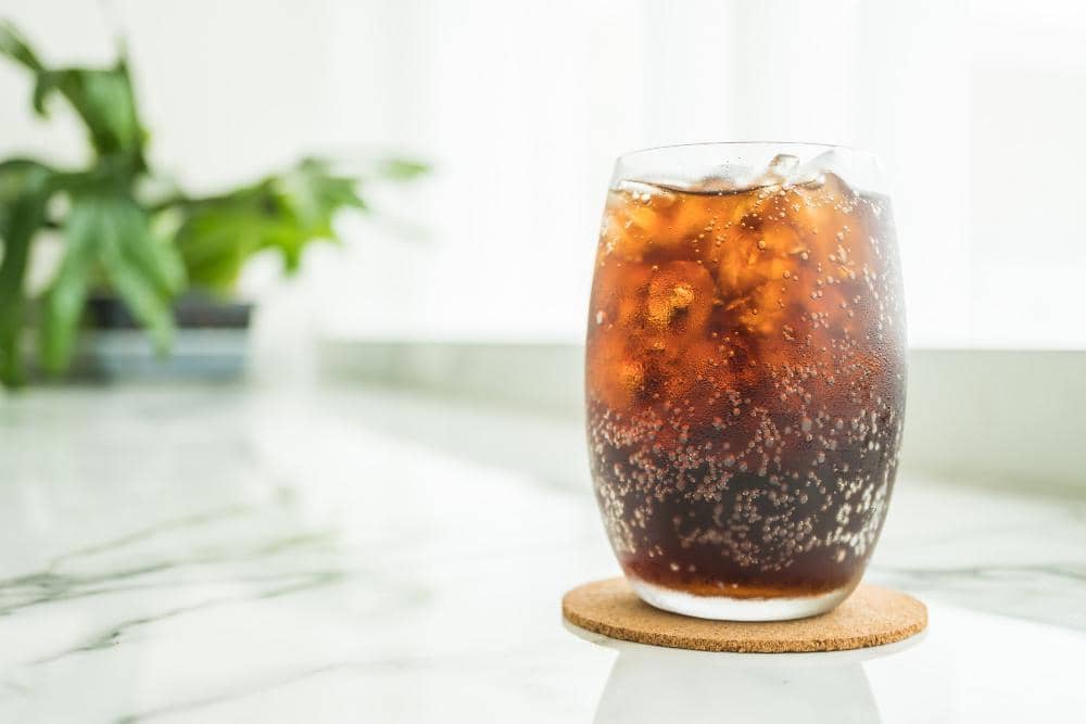 Segelas minuman bersoda berwarna cokelat dengan es batu di atas meja marmer, latar belakang tampak tanaman hijau.
