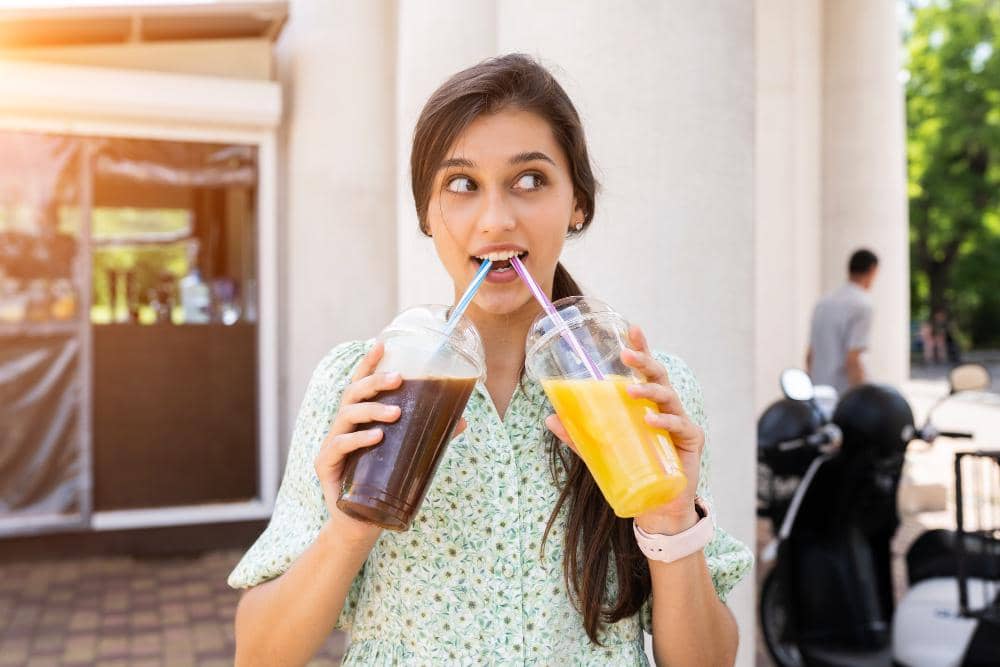 Seorang wanita muda memegang dua gelas plastik berisi minuman berperisa buah, satu berwarna cokelat dan satu berwarna kuning, sambil tersenyum di luar ruangan.