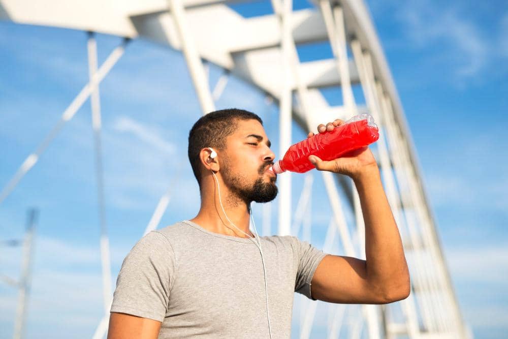 Pria mengenakan kaus abu-abu dan earphone sedang minum minuman energi berwarna merah di luar ruangan dengan latar jembatan putih.