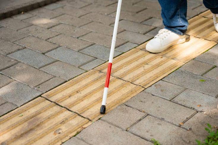 Bagian kaki seseorang yang mengenakan sepatu putih dan celana jin biru sedang melangkah di atas jalur pemandu berwarna kuning. Sebilah tongkat putih dengan ujung merah tampak menyentuh permukaan ubin taktil tersebut untuk mendeteksi jalan.