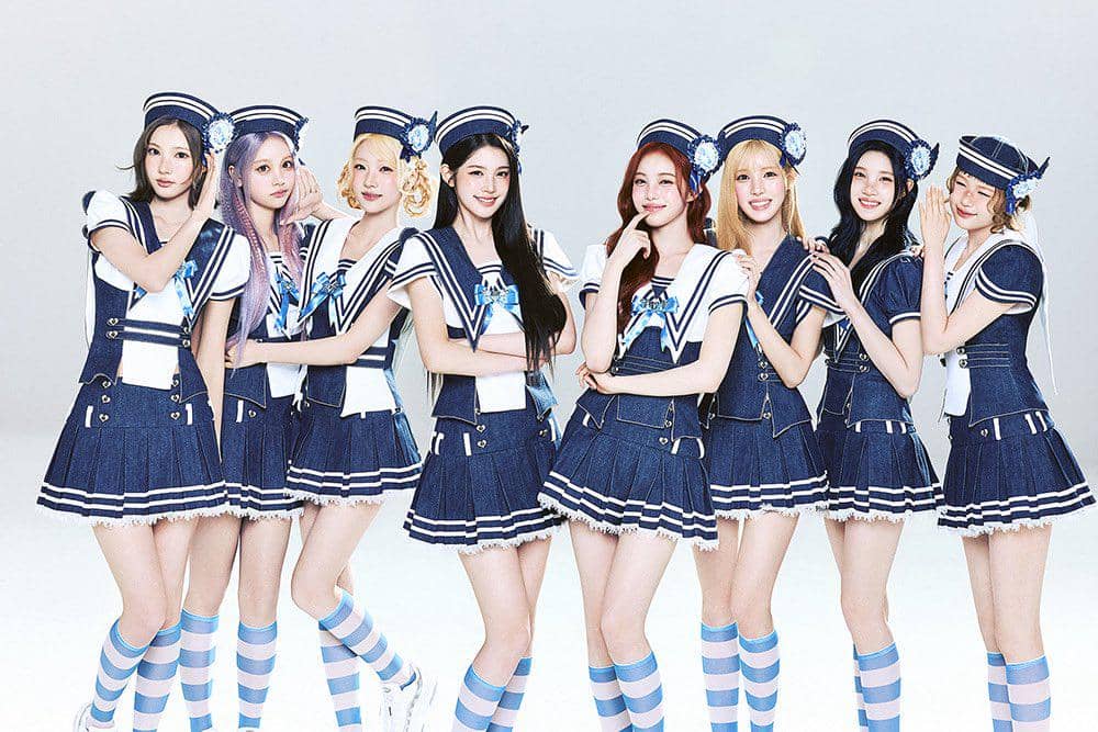 Sembilan perempuan mengenakan kostum bertema pelaut berwarna biru dan putih dengan kaus kaki belang biru muda, berpose bersama di studio.