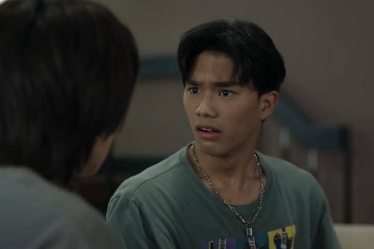 Seorang pria muda mengenakan kaus hijau dan kalung rantai tampak terkejut saat berbicara dengan seseorang di depannya dalam adegan drama WU.