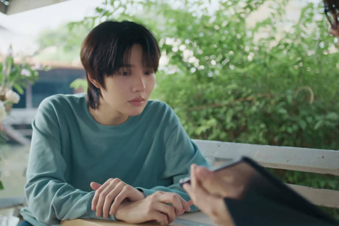 Seorang karakter dalam drama Still Holding up like the Moon duduk di bangku luar ruangan mengenakan sweater hijau muda dan menatap serius ke arah seseorang.