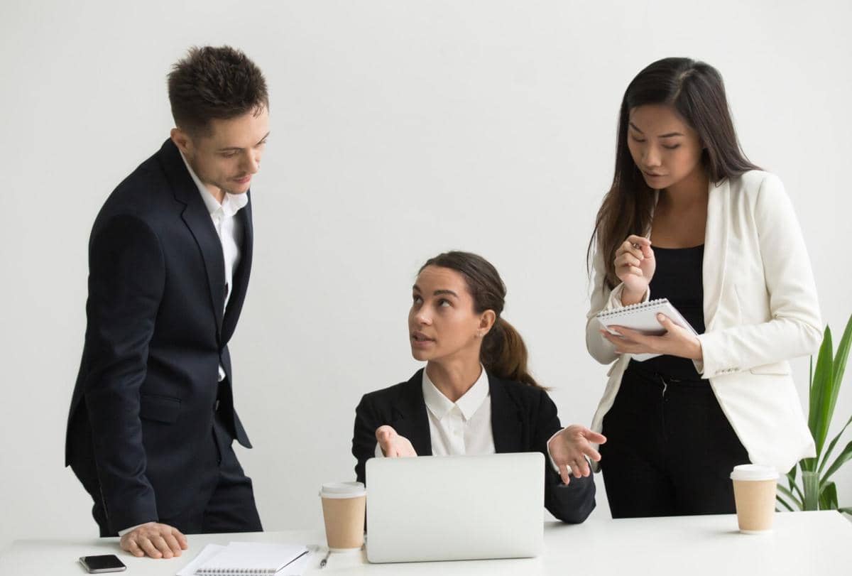 Tiga rekan kerja berdiskusi di kantor, dua berdiri mendengarkan sementara satu duduk di depan laptop dengan ekspresi serius.