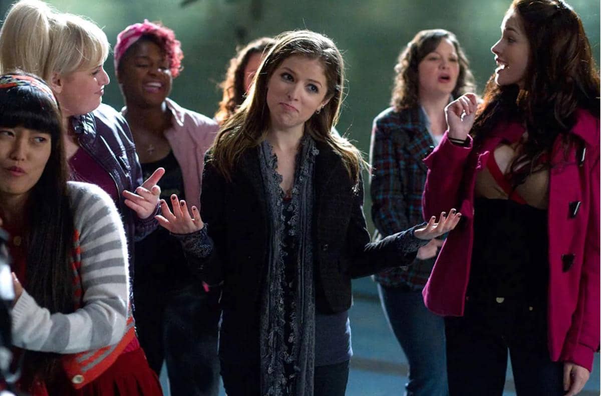 Beberapa perempuan berdiri bersama di luar ruangan, salah satunya mengangkat tangan dengan ekspresi santai dalam adegan film Pitch Perfect.