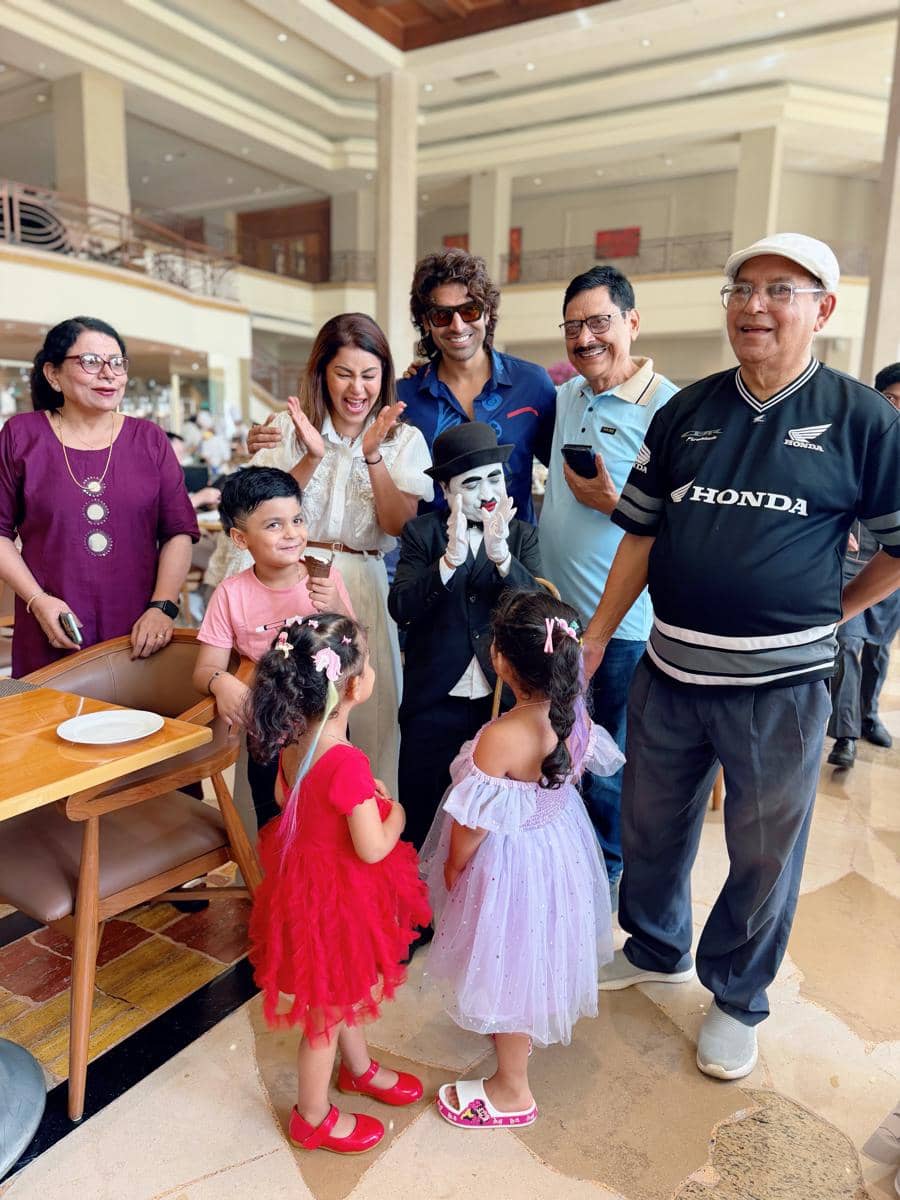 Sekelompok orang dewasa dan anak-anak tersenyum dan tertawa bersama seorang penampil berpakaian seperti Charlie Chaplin di dalam ruangan luas.