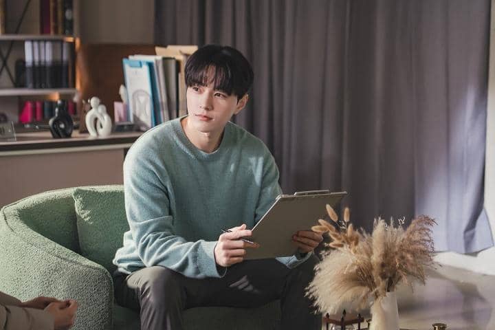 Seorang pria mengenakan sweater biru muda duduk di sofa hijau sambil memegang clipboard di ruangan dengan pencahayaan lembut.