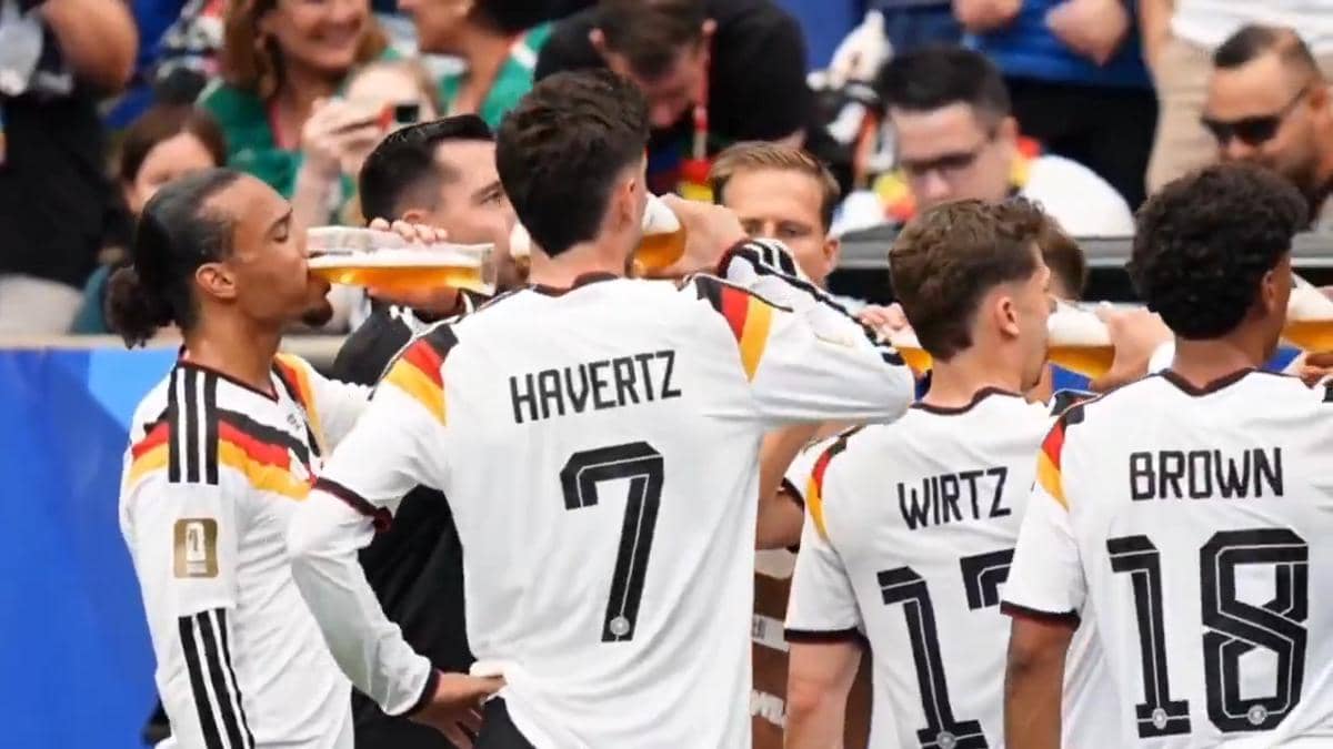 Para pemain tim nasional Jerman mengenakan seragam putih dengan garis hitam, merah, dan kuning sedang minum dari gelas besar di pinggir lapangan.