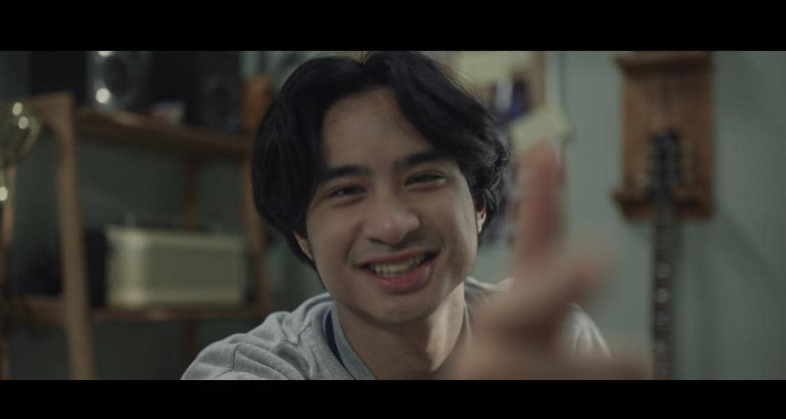Seorang pria muda tersenyum sambil menunjuk ke arah kamera dalam cuplikan film Rumah Singgah dengan latar ruangan sederhana.