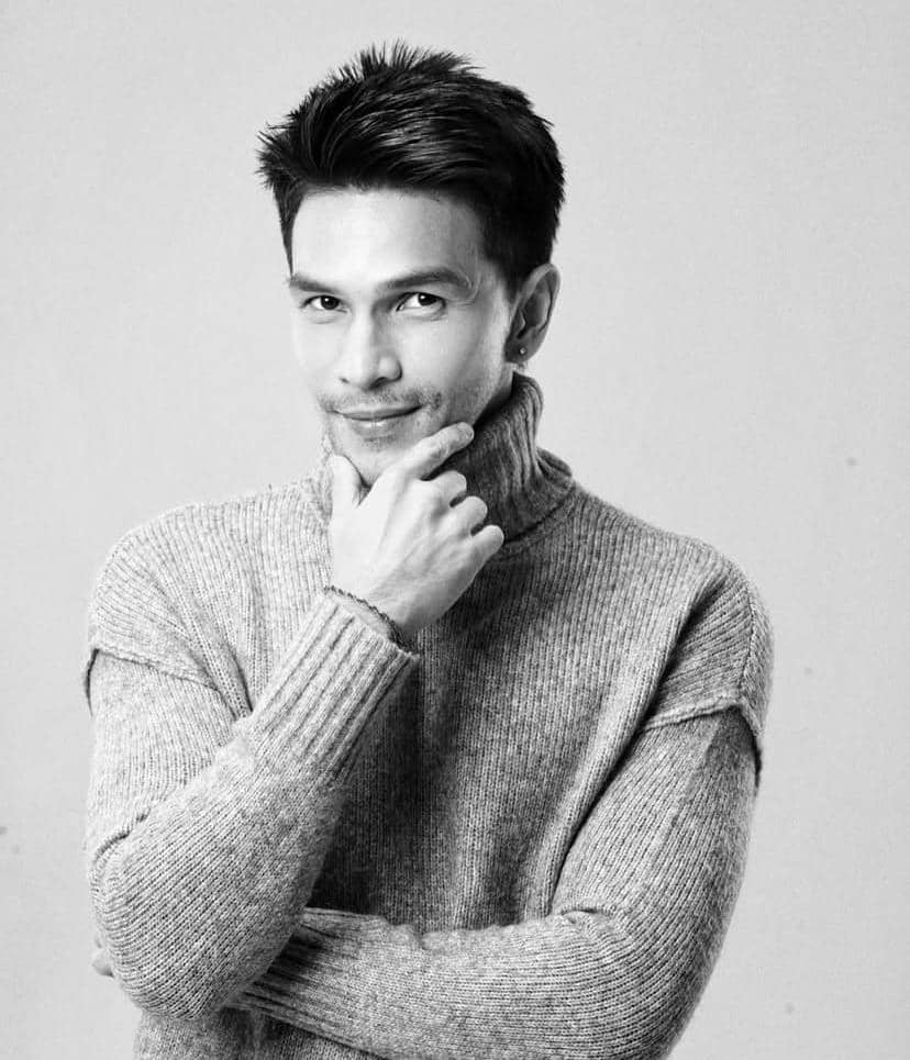 Seorang pria berpose dengan tangan di dagu mengenakan sweater rajut berwarna abu-abu dalam potret hitam putih bergaya studio.