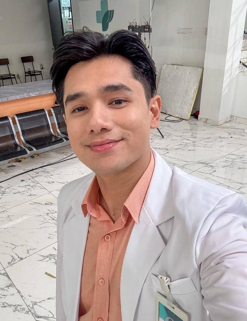 Seorang pria berambut hitam mengenakan kemeja oranye dan jas putih berfoto selfie di ruangan dengan lantai marmer putih.