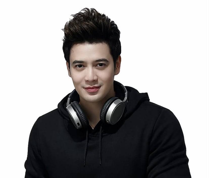 Seorang pria mengenakan hoodie hitam dengan headphone di lehernya, berpose di depan latar belakang putih dengan ekspresi tenang.