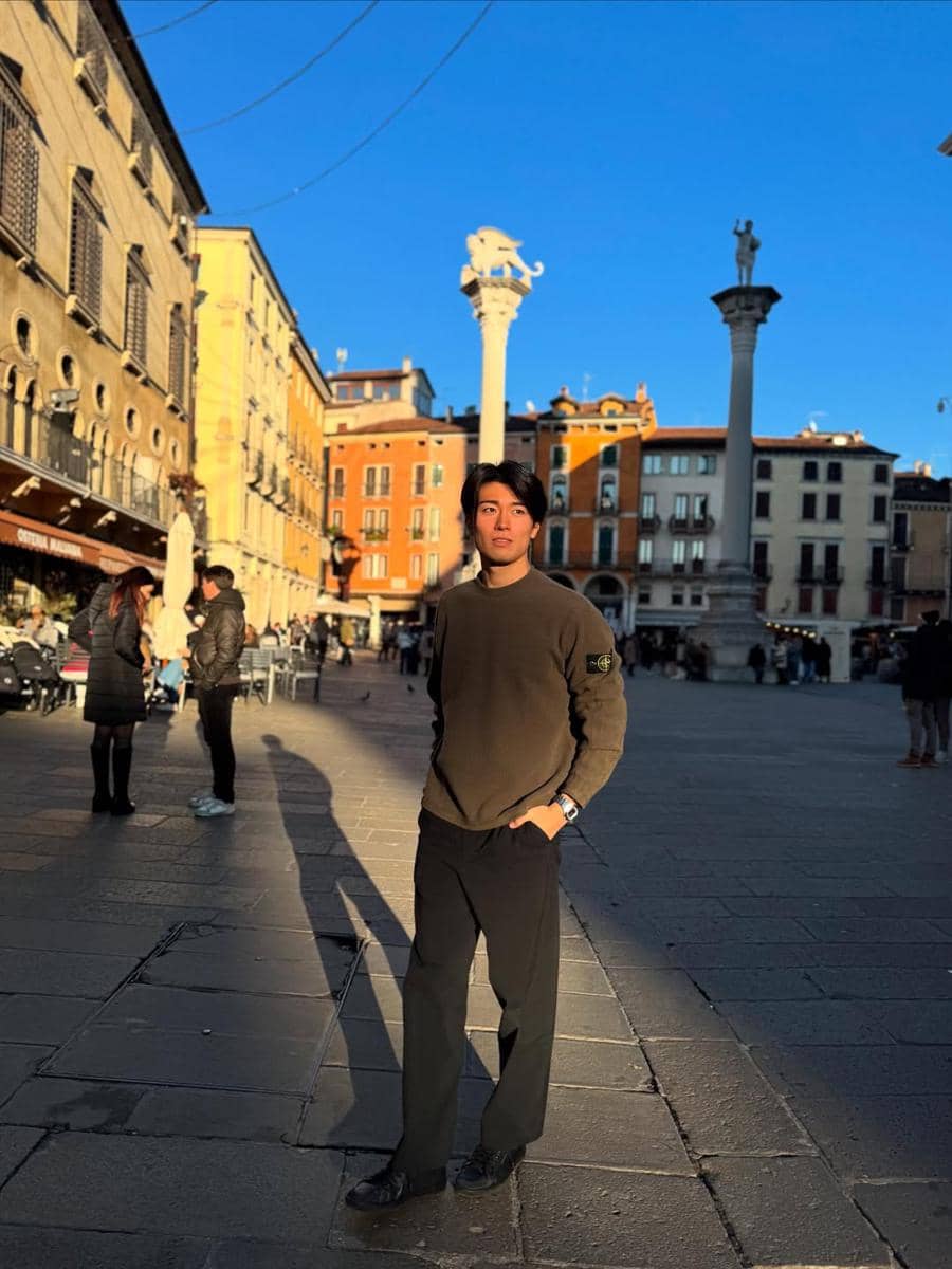 Keito Nakamura berdiri di alun-alun kota dengan latar bangunan bergaya klasik dan dua tiang tinggi di bawah langit biru cerah.