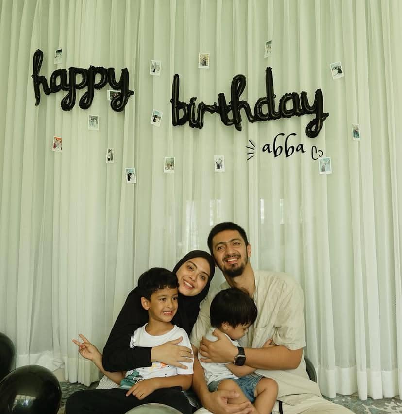 Sebuah keluarga berpose bahagia di depan tirai putih dengan dekorasi ulang tahun bertuliskan happy birthday abba dan balon hitam di sekitarnya.