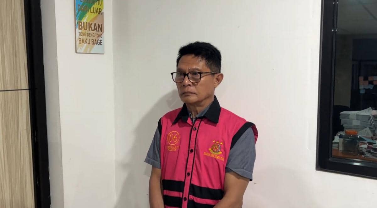 Seorang pria mengenakan rompi tahanan berwarna merah muda dengan logo Kejaksaan, berdiri di dalam ruangan dekat dinding putih.