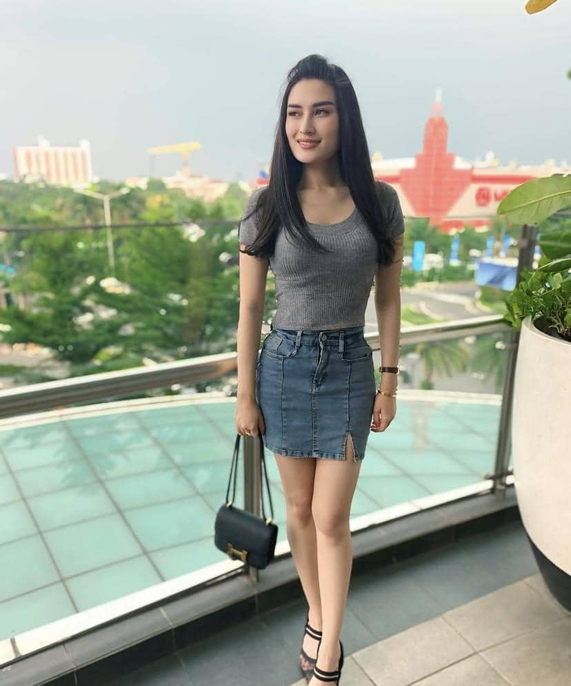 Seorang wanita berambut panjang mengenakan atasan abu-abu dan rok denim berdiri di balkon dengan latar gedung dan pepohonan.