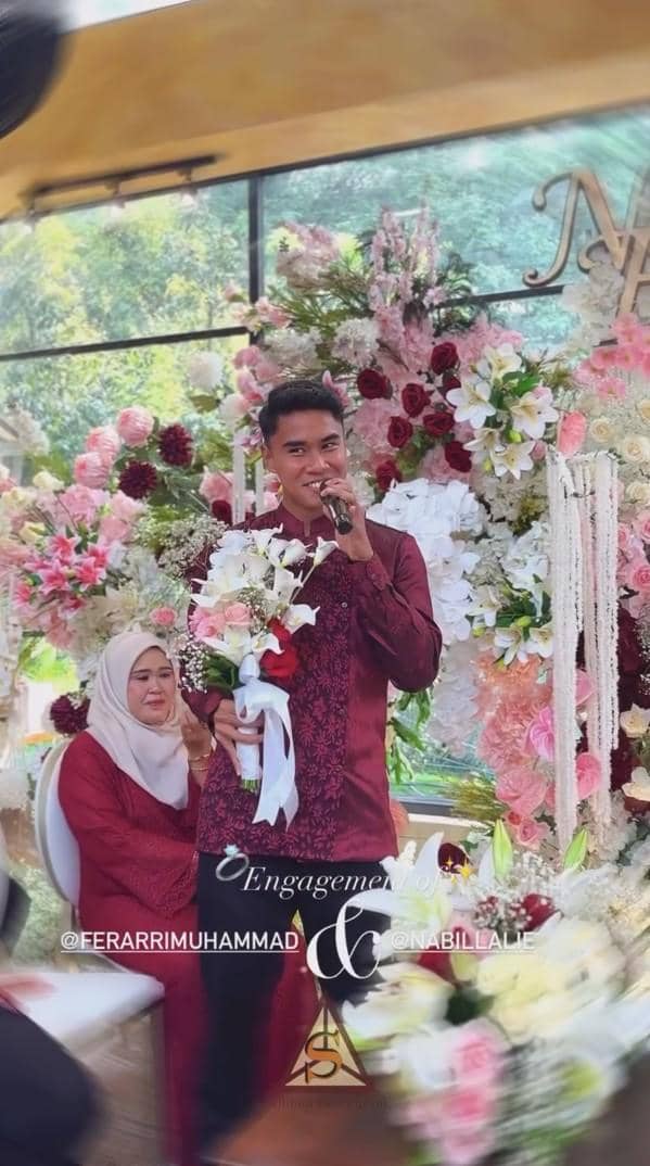 Muhammad Fera mengenakan baju maroon memegang mikrofon dan buket bunga di acara lamaran dengan dekorasi bunga berwarna merah muda dan putih.