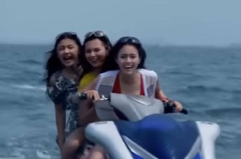 Tiga perempuan muda tertawa bersama saat mengendarai jet ski di laut biru di bawah langit cerah.