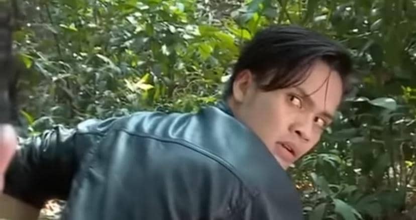 Seorang pria berjaket kulit hitam menoleh ke belakang dengan ekspresi waspada di tengah hutan yang rimbun.