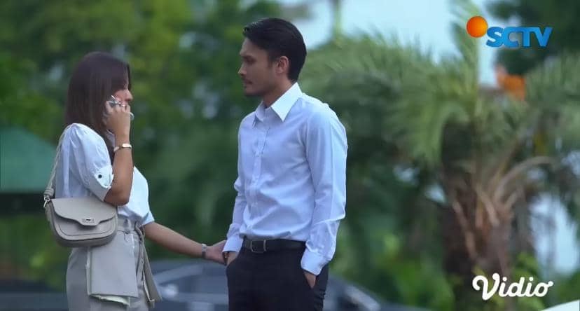 Seorang pria dan wanita berbicara di luar ruangan dengan latar pepohonan, pria mengenakan kemeja putih dan wanita memegang ponsel.