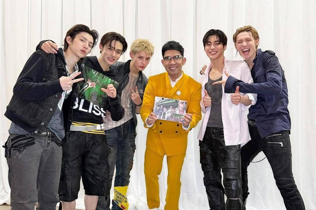 Seorang pria mengenakan setelan kuning berfoto bersama lima pria muda berpakaian kasual di depan latar putih sambil memegang album.