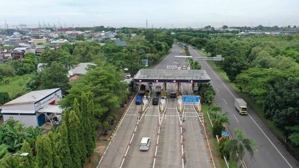 Gerbang tol Makassar dengan beberapa kendaraan melintas di jalur masuk dan keluar, dikelilingi pepohonan hijau dan area permukiman.