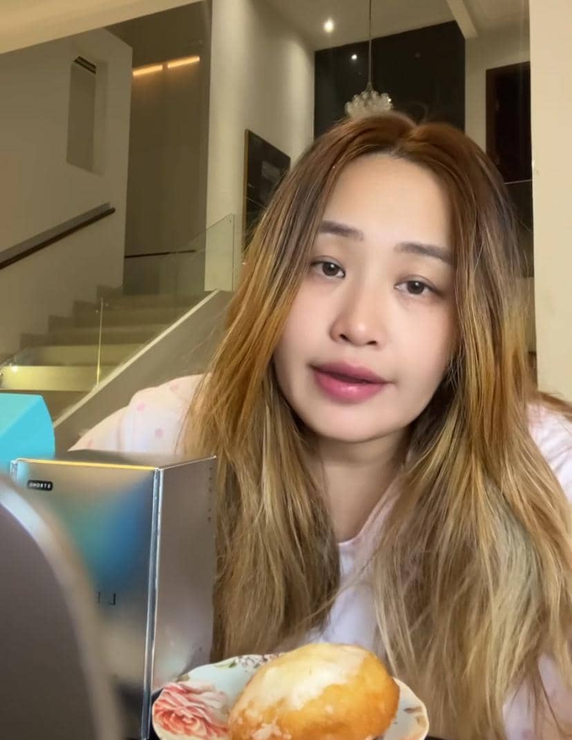 Seorang perempuan berambut panjang pirang duduk di dalam rumah dengan pencahayaan hangat, di depannya terdapat makanan dan cermin kecil.