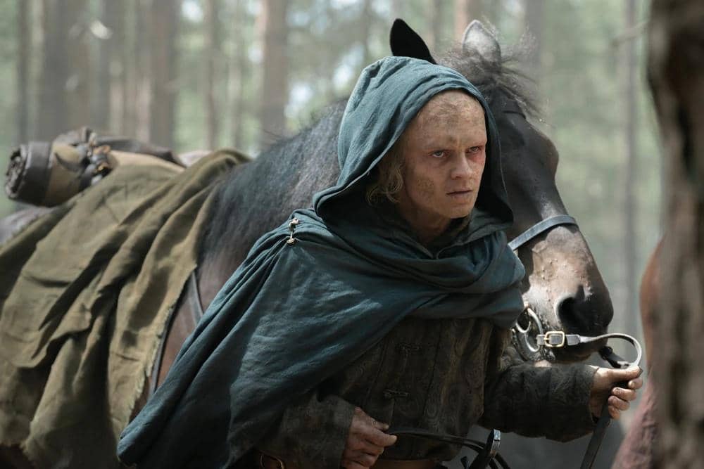 Karakter Aegon II mengenakan jubah berkerudung hijau tua sambil memegang tali kekang kuda di hutan dalam adegan House of the Dragon Season 3.
