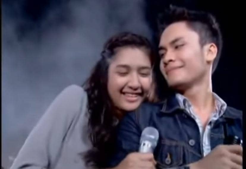 Dua remaja tersenyum sambil memegang mikrofon di panggung dengan latar asap tipis, menunjukkan suasana romantis dan bahagia.